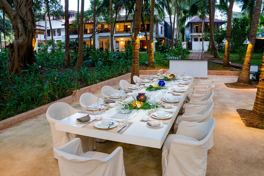 A long table laid for dinner under the palms, the lit house behind it