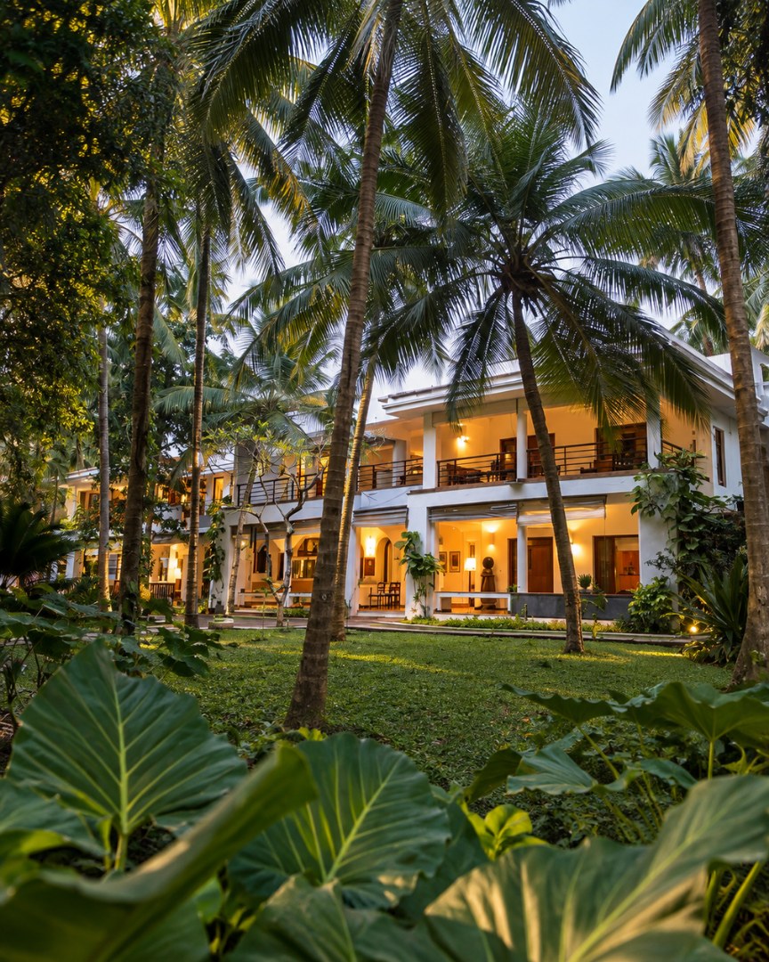 Villa Aashyana at dusk, lit from within, seen across the lawn through coconut palms